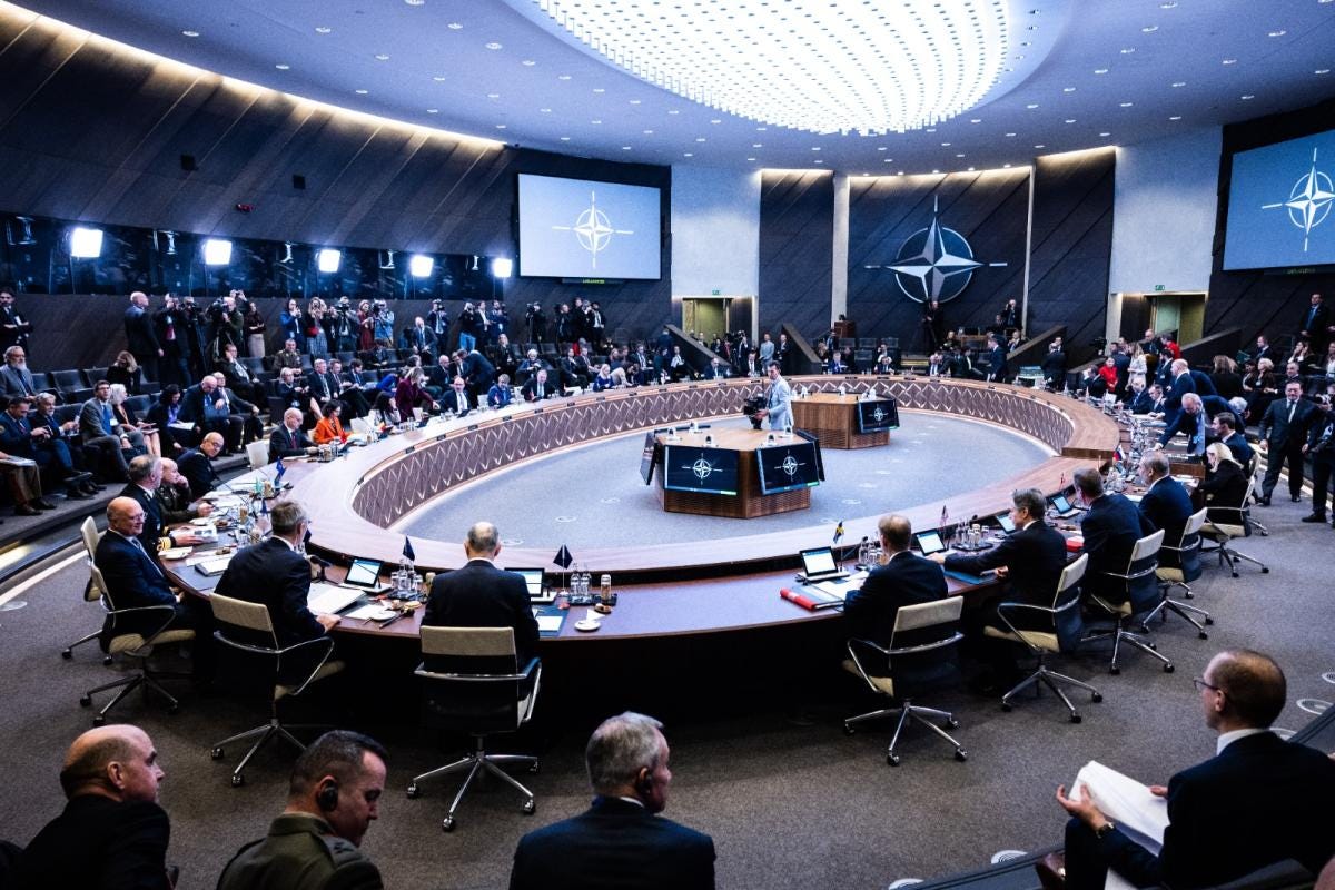 Photo of the NATO meeting room. Around an oval table are numerous people. In the background the logo of NATO