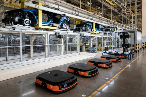 In a large industrial setting, a row of low-slung wheeled vehicles sits beside several vehicles under construction. 