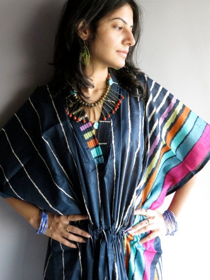 May include: A navy blue kaftan dress with white vertical stripes and colorful accents. The dress features a V-neckline with a decorative panel of turquoise, purple, and orange. The sleeves have a striped pattern with pink, orange, and teal. The dress is cinched at the waist with a drawstring.