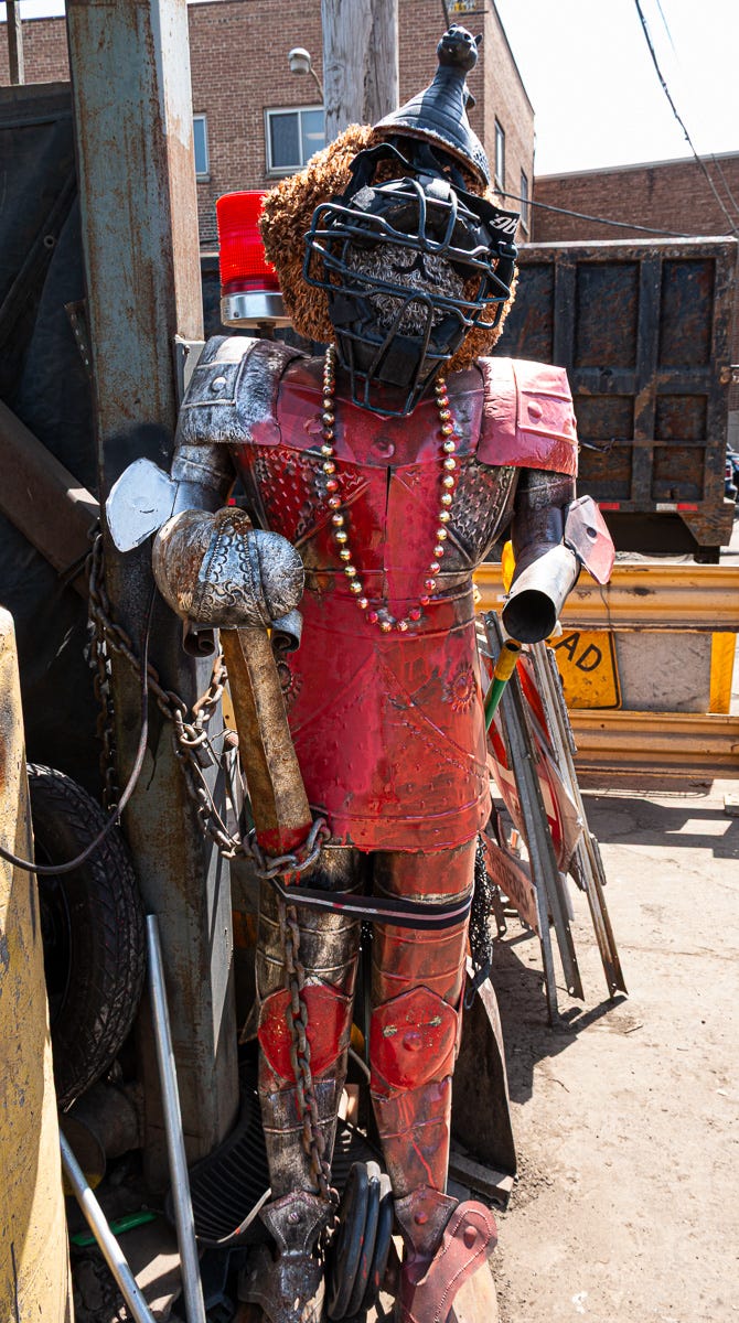 A close-up of a humanoid scarecrow-like figure built from scrap metal, posed among industrial debris and rusted components. A close-up of a humanoid scarecrow-like figure built from scrap metal, posed among industrial debris and rusted components.