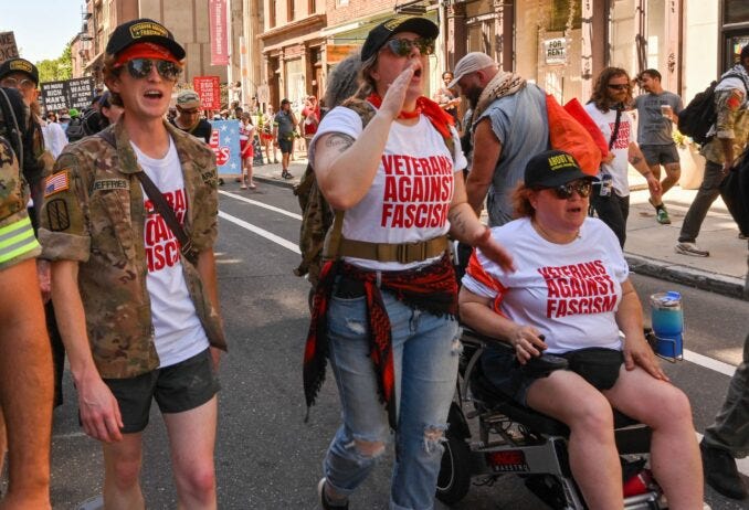 Fotografía de tres mujeres veteranas desfilando en Filadelfia el 4 de julio. Llevan camisetas que dicen « VETERANS AGAINST FASCISM ». Una de las mujeres está en silla de ruedas, las otras dos caminan a su lado. Se ven decididas y unidas contra el fascismo, celebrando a su manera el Día de la Independencia. La escena transcurre en una calle soleada de la ciudad de Filadelfia, con una bandera usamericana visible al fondo. Fotografía de tres mujeres veteranas desfilando en Filadelfia el 4 de julio. Llevan camisetas que dicen « VETERANS AGAINST FASCISM ». Una de las mujeres está en silla de ruedas, las otras dos caminan a su lado. Se ven decididas y unidas contra el fascismo, celebrando a su manera el Día de la Independencia. La escena transcurre en una calle soleada de la ciudad de Filadelfia, con una bandera usamericana visible al fondo.