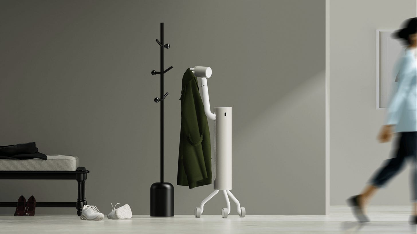 Nome, a domestic robot designed by Frog. A tall, cylindrical white robot with a single articulated arm rolls on four small wheels in the entryway of a home. It is lifting a green jacket onto a black wooden coat rack, next to a bench with a black cushion, a pair of white sneakers, and red shoes on the floor. In the background, a person walks past a picture frame on the wall. The scene is muted, minimalist, and warmly lit.