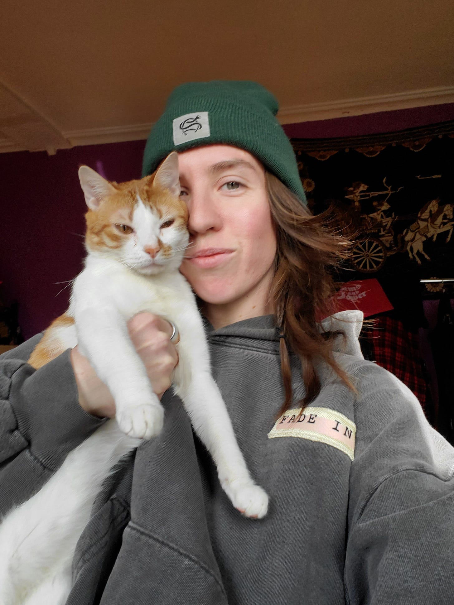 The author, a white transmasculine person with long reddish brown hair sneaking out of a green beanie, snuggles an orange and white cat to their face, proudly displaying him before the camera.