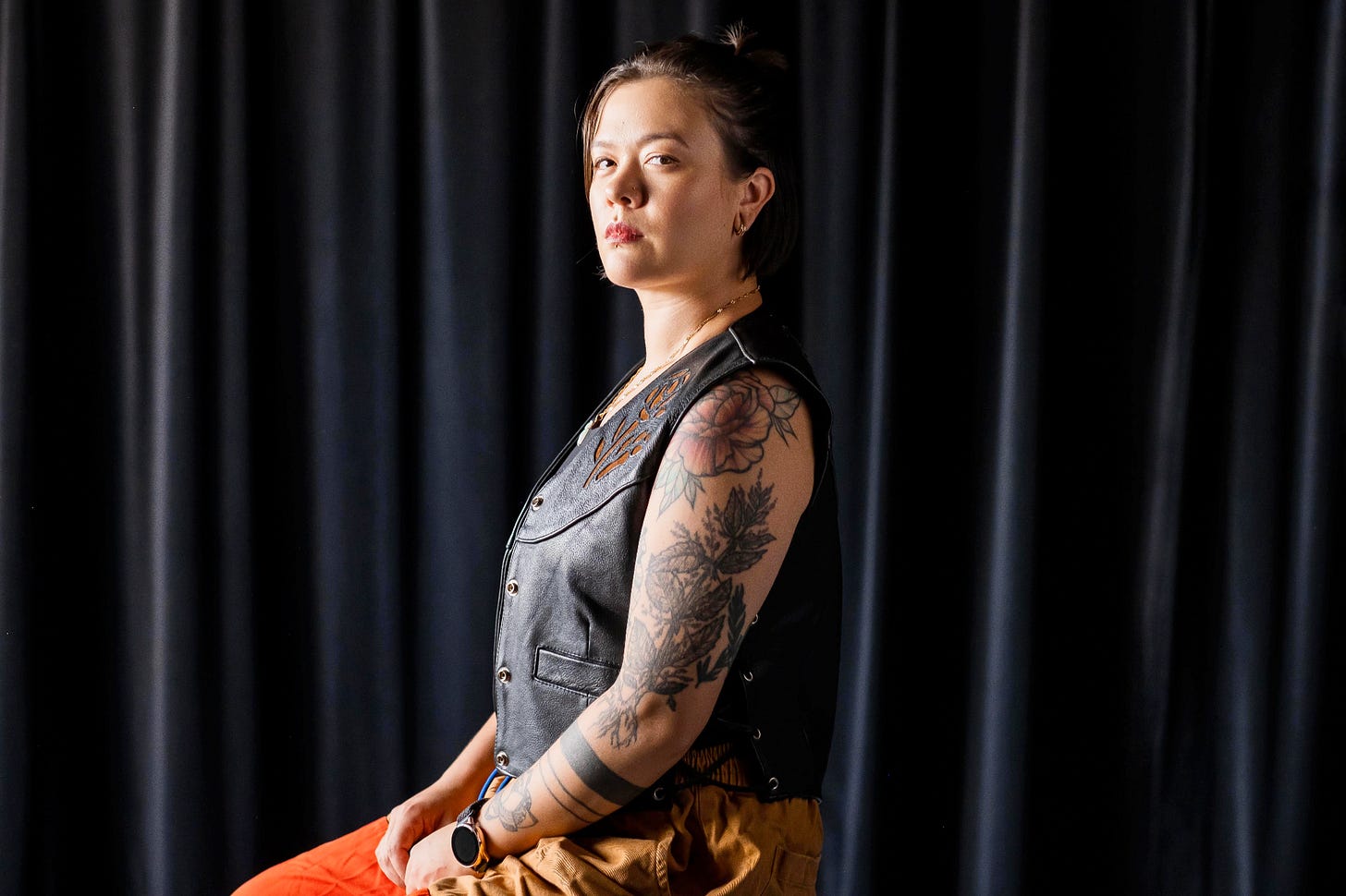 a nb East Asian with short hair and tats wearing a vest gazes at the viewer in profile