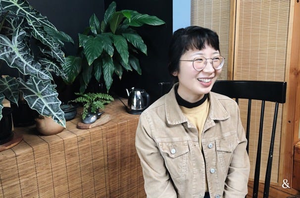 Tiffany in a brown jacket smiling sitting in front of houseplants.
