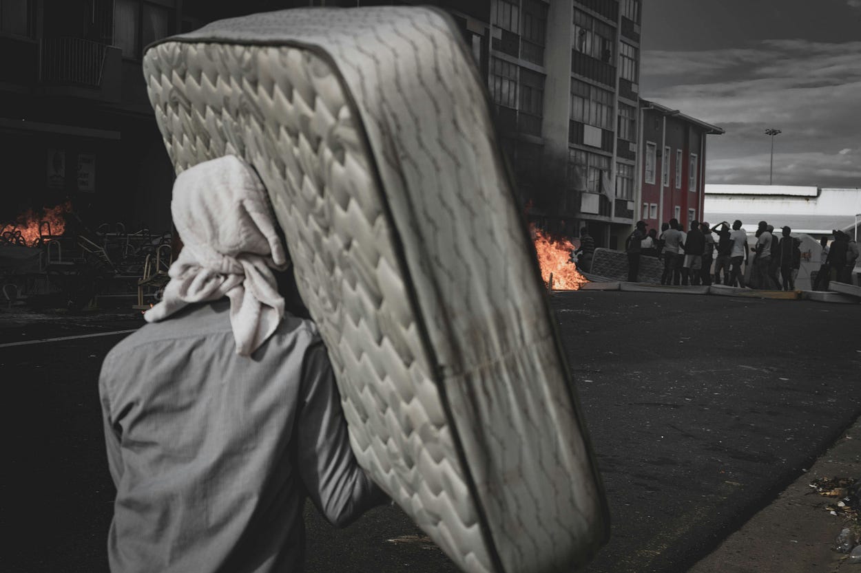 A man dressed in bedraggled clothing carries a battered mattress toward a building. There is a line of people outside the building and a large fire.