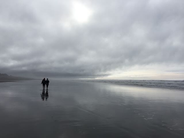 Two people who are clearly close friends walk next to the ocean and the sand looks like a mirror as the water has spread over it