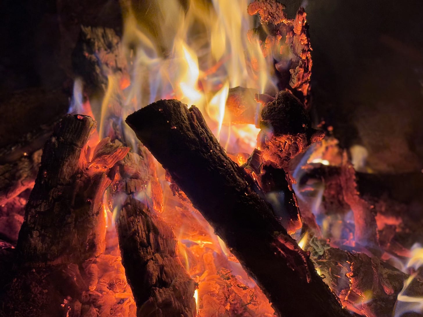 A close-up of a campfire at night highlights the glowing logs in sharp contrast, while the yellow, orange and purple of the flames is slightly blurred with an almost liquid feel. A close-up of a campfire at night highlights the glowing logs in sharp contrast, while the yellow, orange and purple of the flames is slightly blurred with an almost liquid feel.