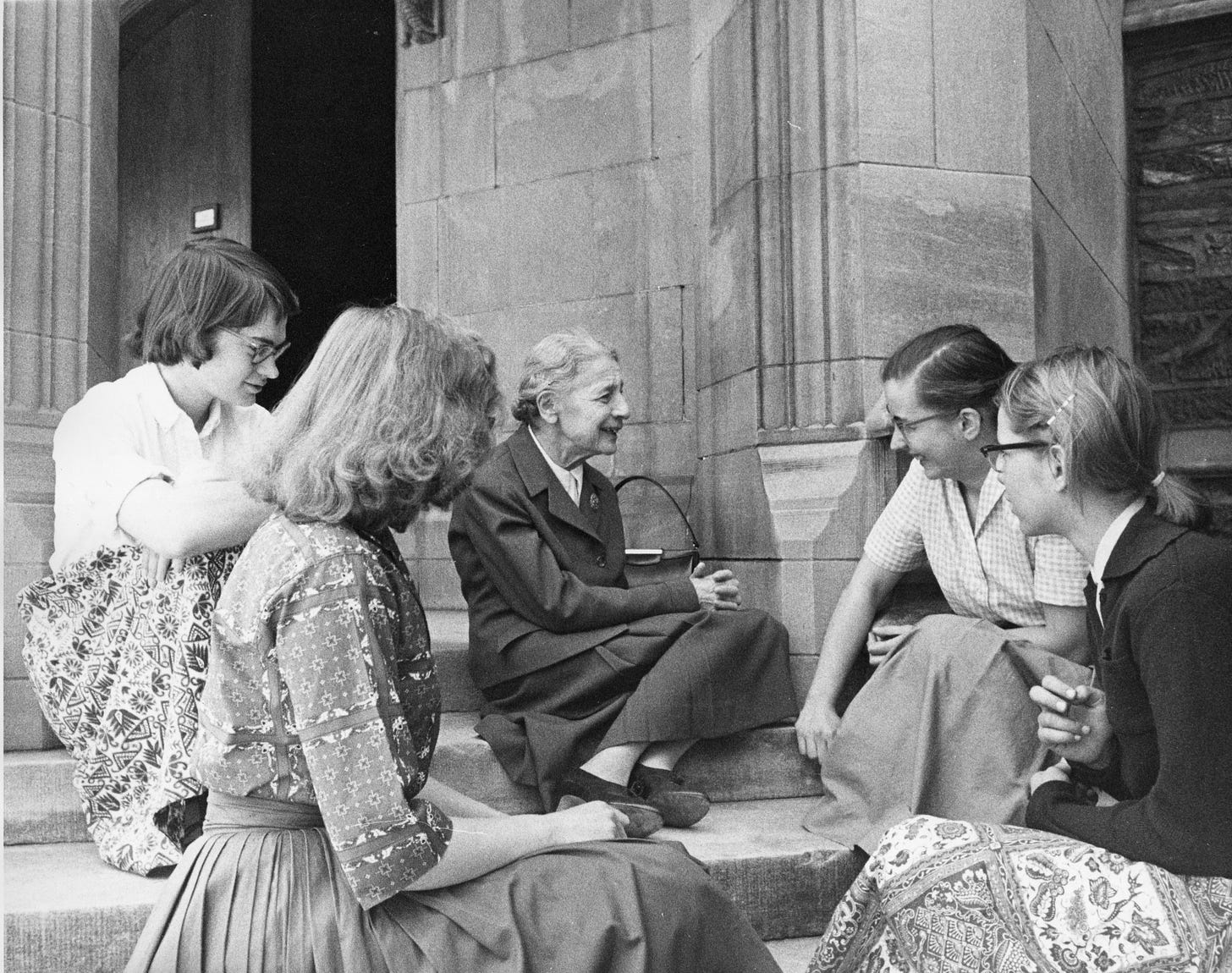BLack and white photo of students (all women) sitting on the steps of a building listening to an older woman talk
