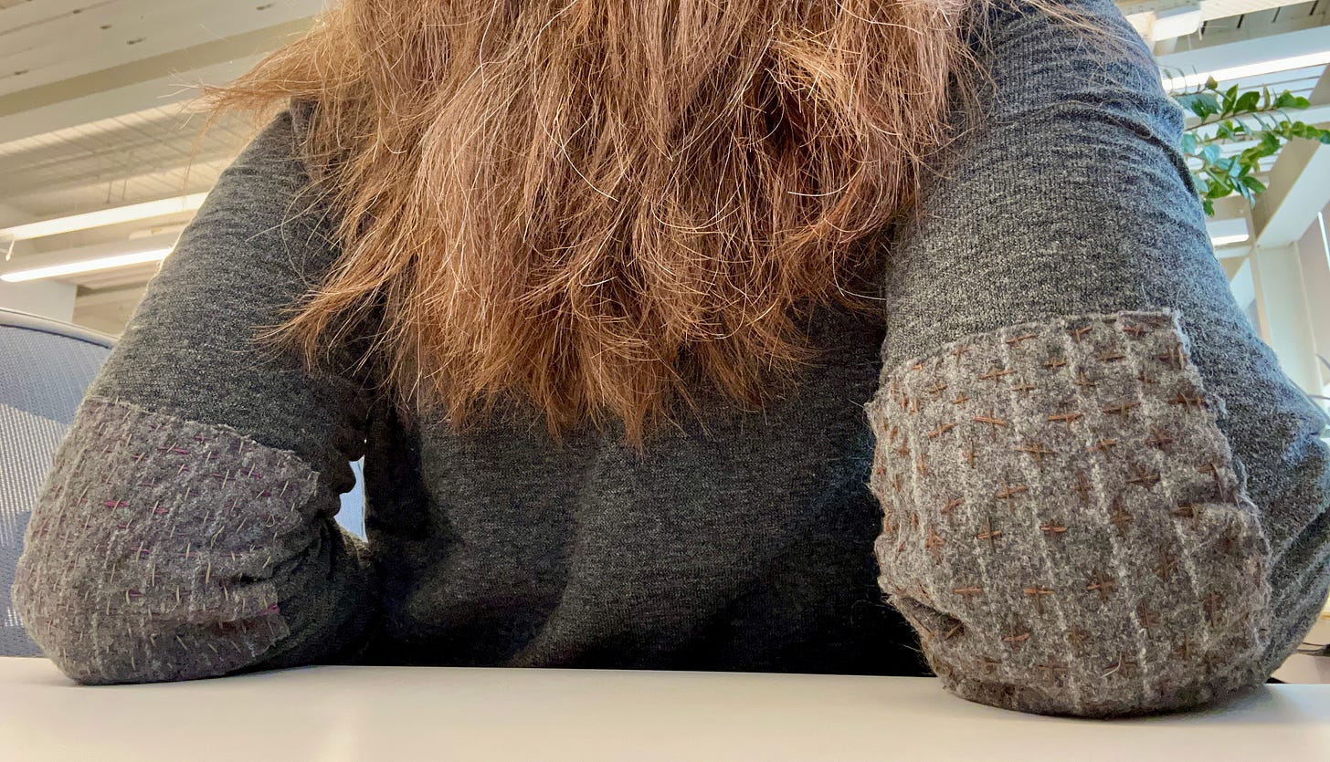 woman's back showing brownish red hair and grey sweater with patched elbows