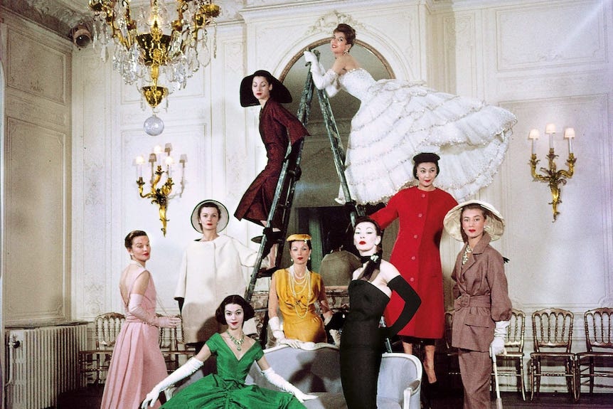 Women in dresses stand around a couch and a ladder while posing for a photo Women in dresses stand around a couch and a ladder while posing for a photo