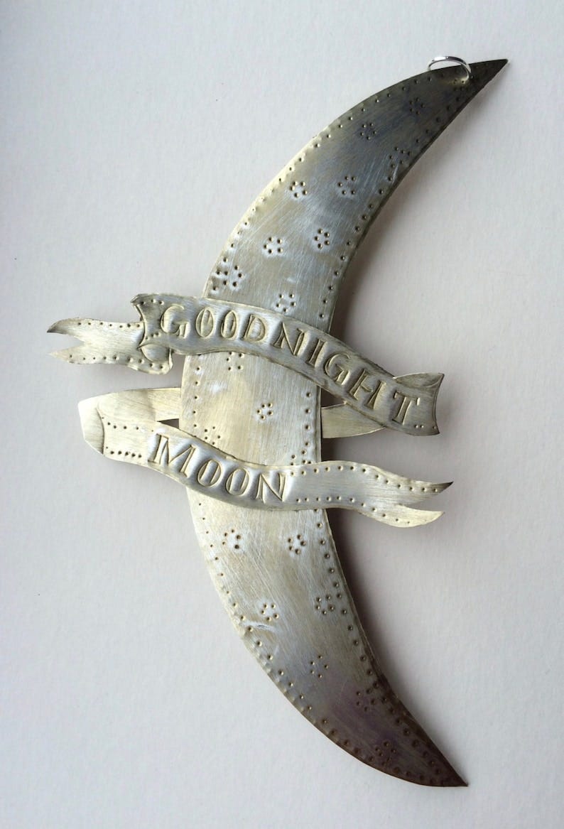 May include: A silver metal crescent moon with the words 'Good Night Moon' embossed on it. The moon has a small loop at the top for hanging.