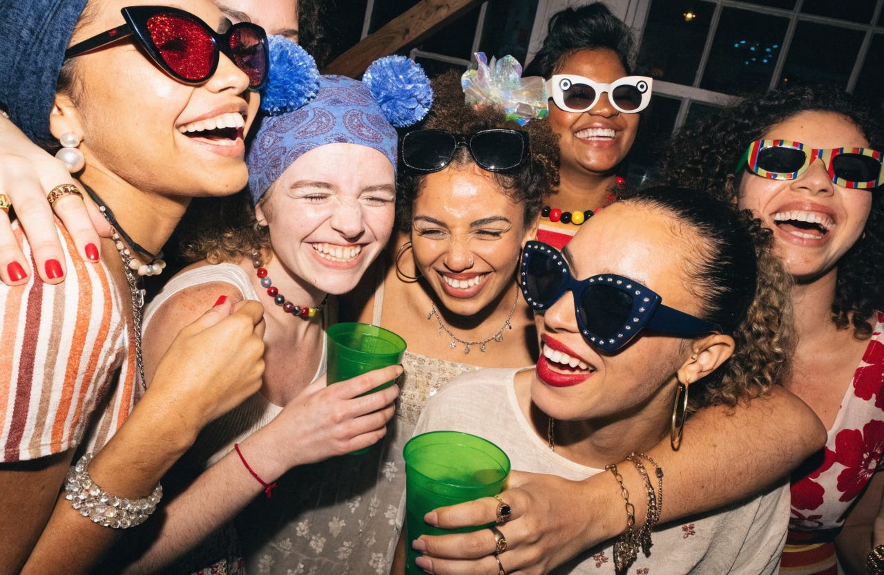 A group of young women friends, mixed ethnicities, wearing short party dresses, smiling and laughing with their arms around each other at a house party in Shoreditch, London.
