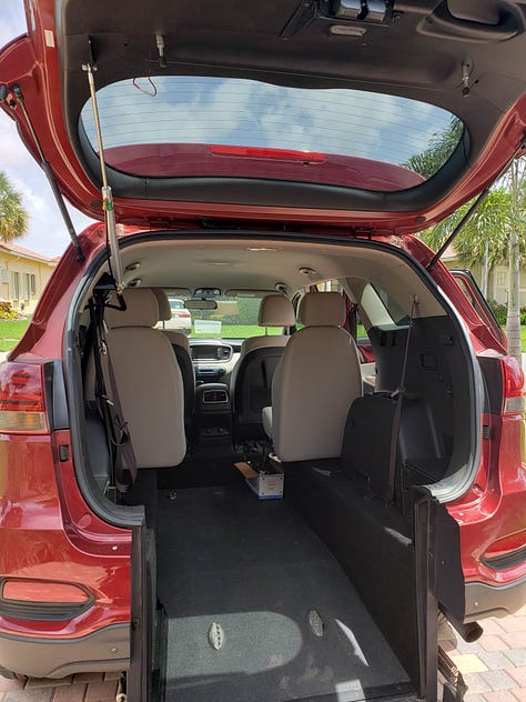 There are 3 photos in a gallery. From left to right: the first photo is the back of a white SUV, but it has been modified and is higher off the ground than normal; the trunk is open, and a wheelchair ramp extends out the back onto the ground. In the second, a red SUV similar to the first in that it has been modified; this view shows the interior of the back, with a built-in wheelchair ramp. The third is a picture of the writer of this piece; she is standing in a driveway with a walker, wearing a burgundy cardigan, black tank top, jeans, one sneaker, and one boot on her feet; behind her is a Silver 2023 Honda Odyssey minivan.