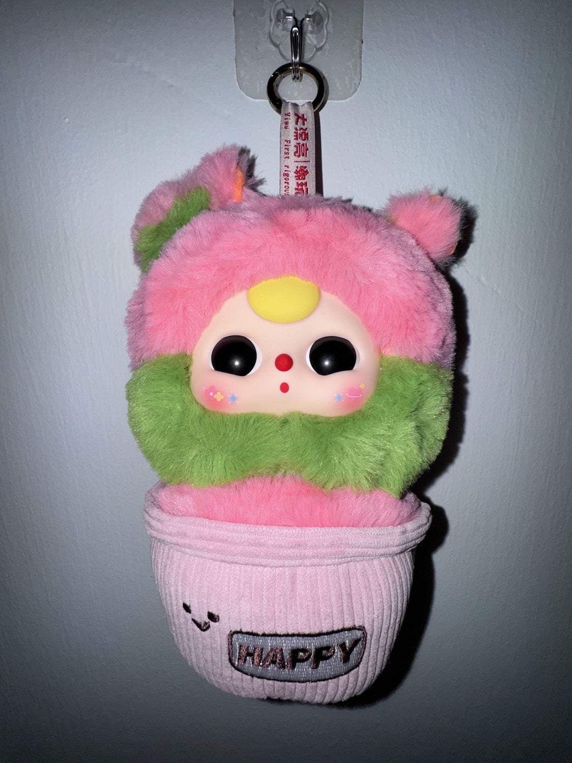 Small Baby Three plush keychain hanging on a wall: pink fur with a green collar, yellow forehead dot, black button eyes, sitting in a ribbed pink fabric pot embroidered “HAPPY,” suspended from a hook by a fabric loop. Small Baby Three plush keychain hanging on a wall: pink fur with a green collar, yellow forehead dot, black button eyes, sitting in a ribbed pink fabric pot embroidered “HAPPY,” suspended from a hook by a fabric loop.