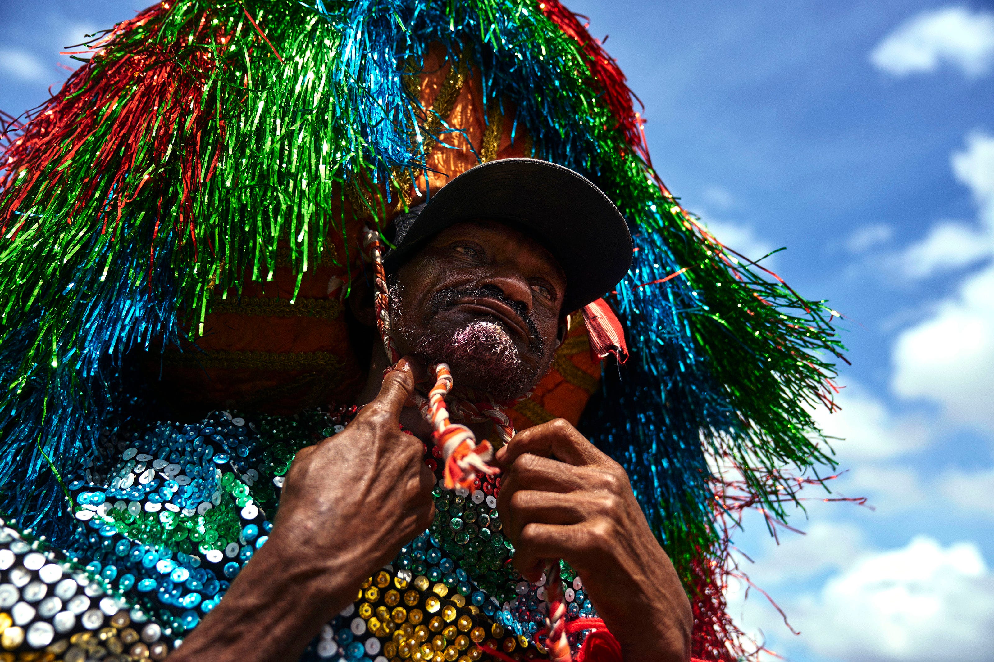A person wearing a colorful, sequined costume and a headdress made of shiny tinsel strands in green, blue, and red, looks upward against a bright blue sky with scattered clouds.