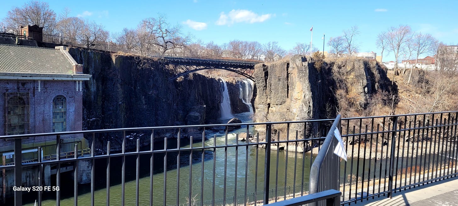 A waterfall. There is a bridge over it, a stone building on the left side, and a railing in the foreground of the picture.