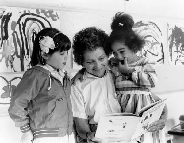 Black and white photograph of Vivian Gussin Paley, a woman with curly dark hair, reading a book with two young girls in a classroom. One girl wears a jacket with a large white bow in her hair, while the other wears a striped dress. Children's artwork is visible on the wall behind them.