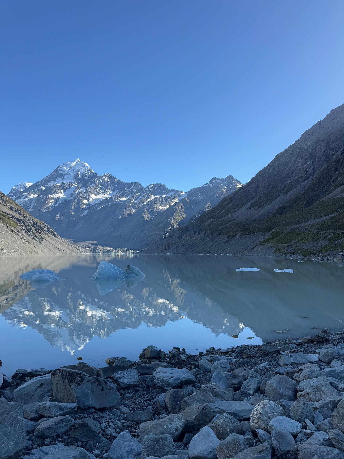 Aoraki-Mt Cook sits against a clear blue sky in the distance, reflected in the water of a glacial lake beneath