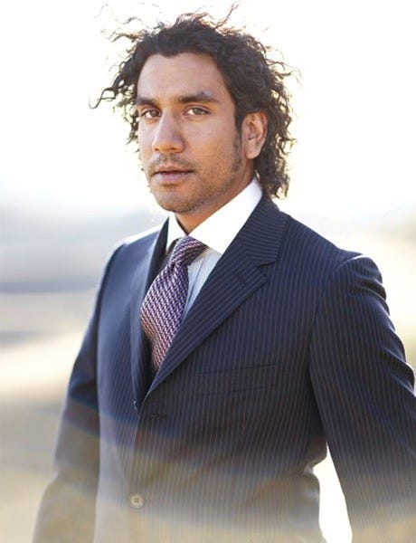 Full body shot of Naveen Andrews in a blue suit and checkered tie in front of a blurred background
