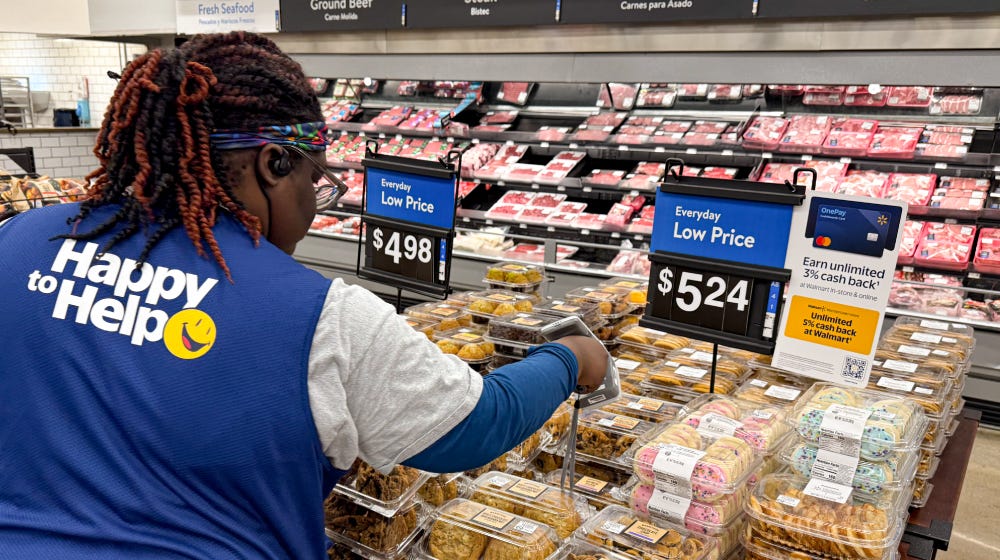 An associate wearing a blue vest with 'Happy to Help' text is organizing packaged food items on a store shelf. The setting is a grocery store with visible signage indicating prices, including '$4.98'. The background displays a meat section with various products.