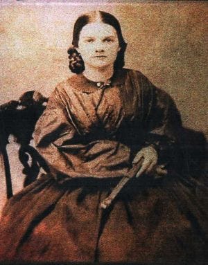 Photo from mid to late 1800s of a young white woman in a black dress that looks too big for her. She is seated on a chair and holding a long object. Her hair is combed back from her face and she has a serious expression. Photo from mid to late 1800s of a young white woman in a black dress that looks too big for her. She is seated on a chair and holding a long object. Her hair is combed back from her face and she has a serious expression.