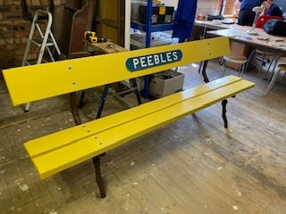 a bright yellow wooden bench with Peebles on a metal sign sits in the Quadrange off Peebles high street near the war memorial.