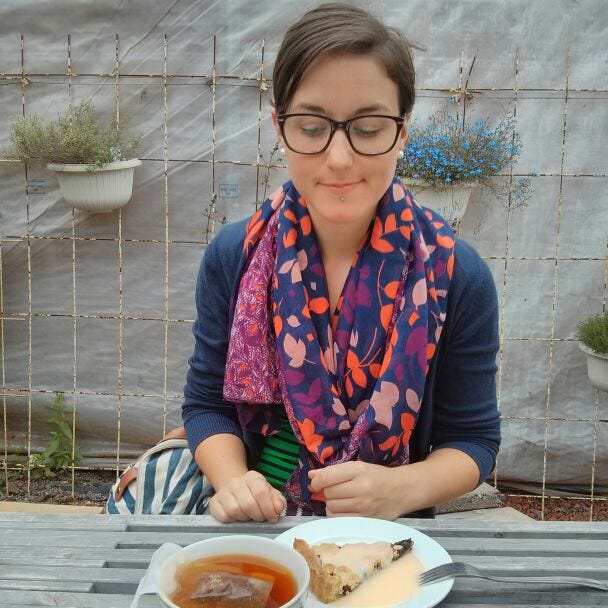 A white woman with short brown hair and glasses is wearing a navy sweater and a colorful scarf. She is looking at and sitting in front of a piece of cake covered in vanilla sauce and a cup of tea. There are plants visible in the background.