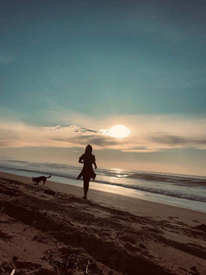 A long, distinct shadow of a person stretches across a wide, flat sandy beach, with a low dune line topped with sparse green and dead trees, and a small full moon visible in the clear blue sky.