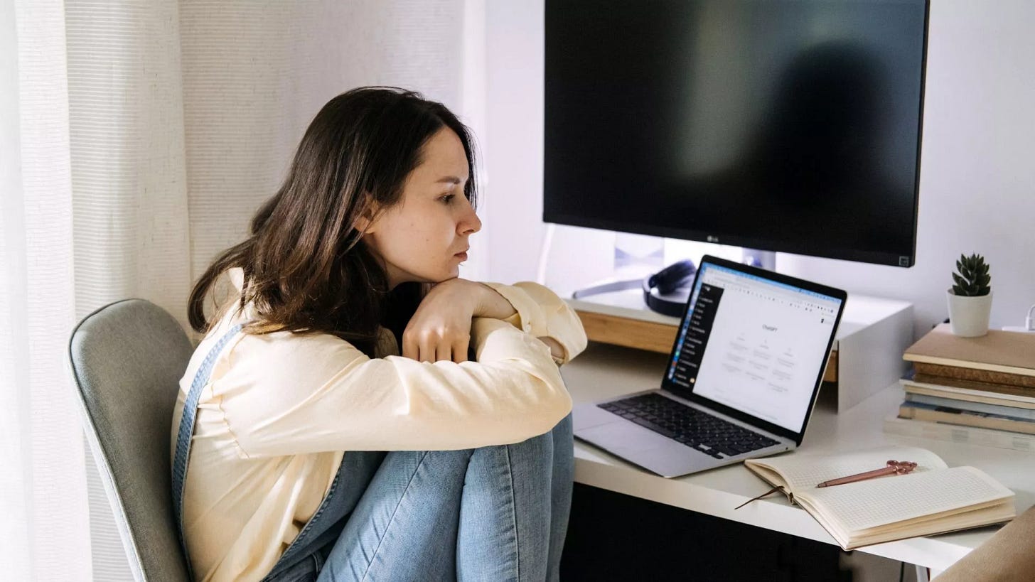 Young woman at desk looking thoughtfully at a laptop with notebook and monitor nearby.