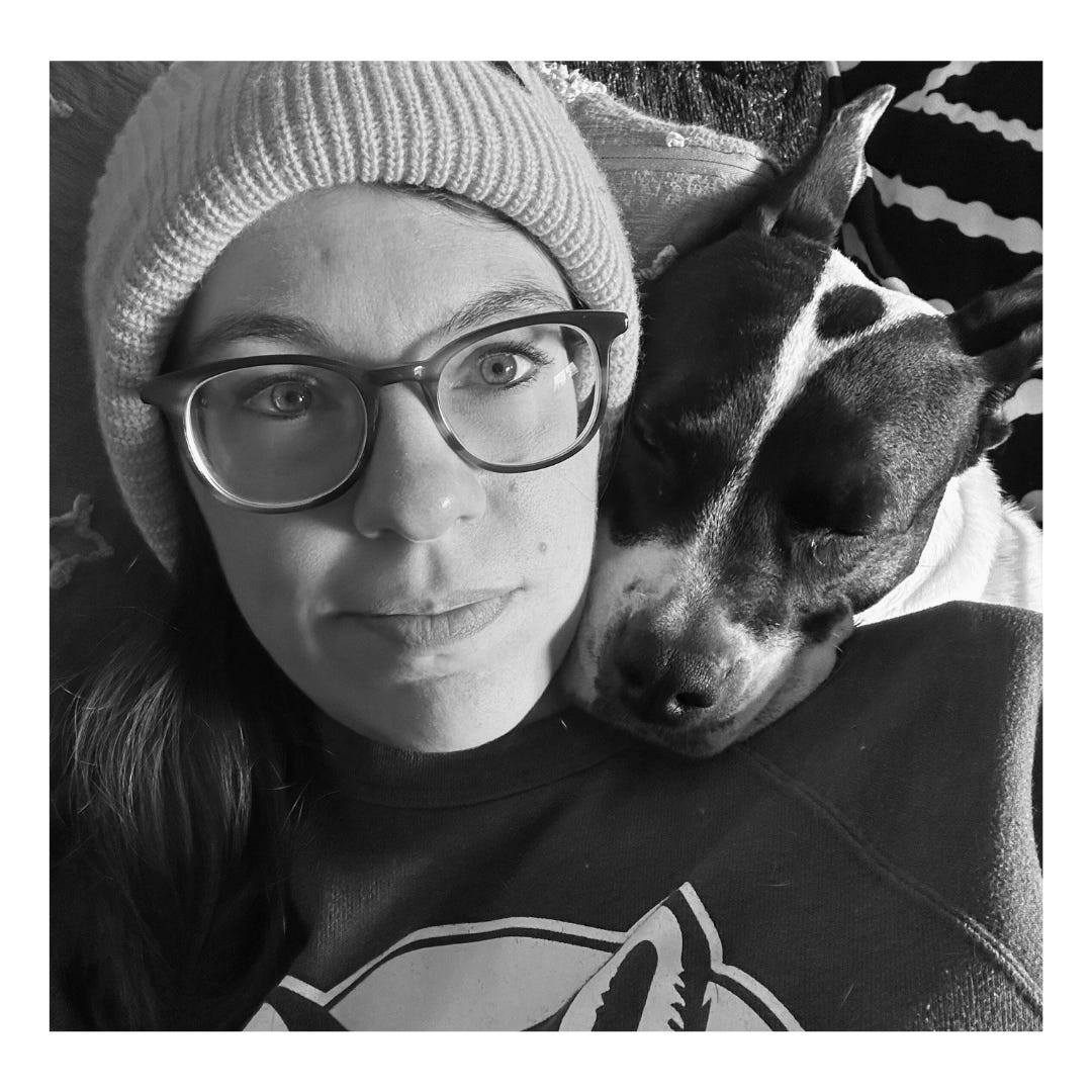 Black and white photo showing a white woman with glasses and a knit cap, laying next to a black and white pitbull who appears to be sleeping on her shoulder.