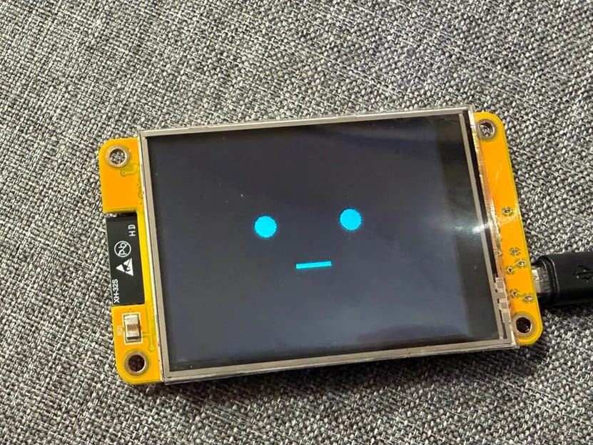 Photo of a yellow ESP32 touchscreen on a gray fabric surface displaying a minimal blue digital face with two round eyes and a straight-line mouth on a dark screen.