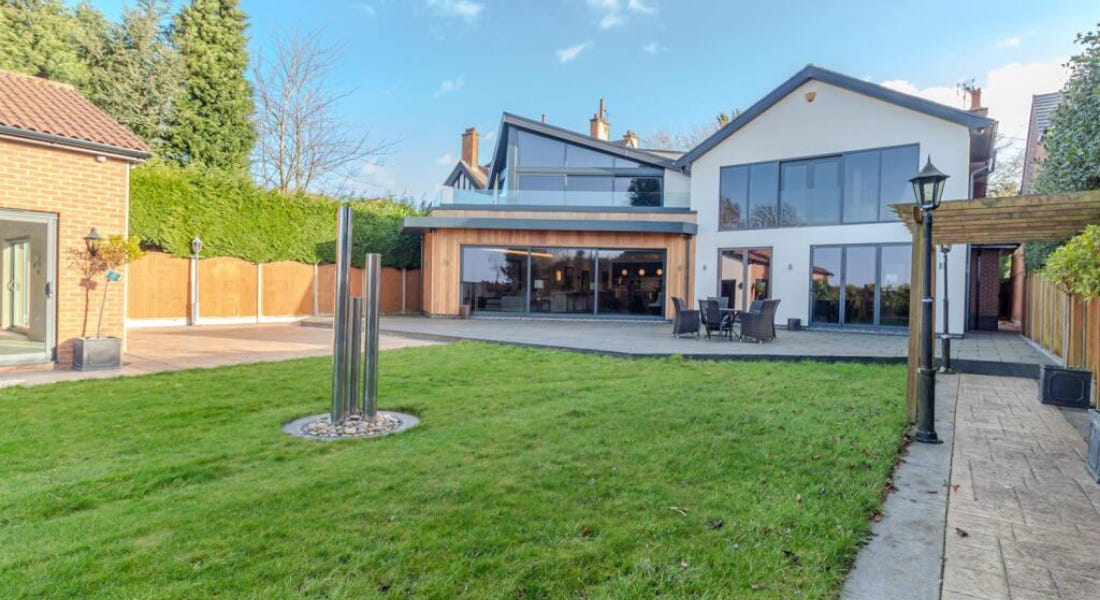 A backyard with a green lawn with umbrella poles, a patio with grey dining seats, and a white and wooden house 