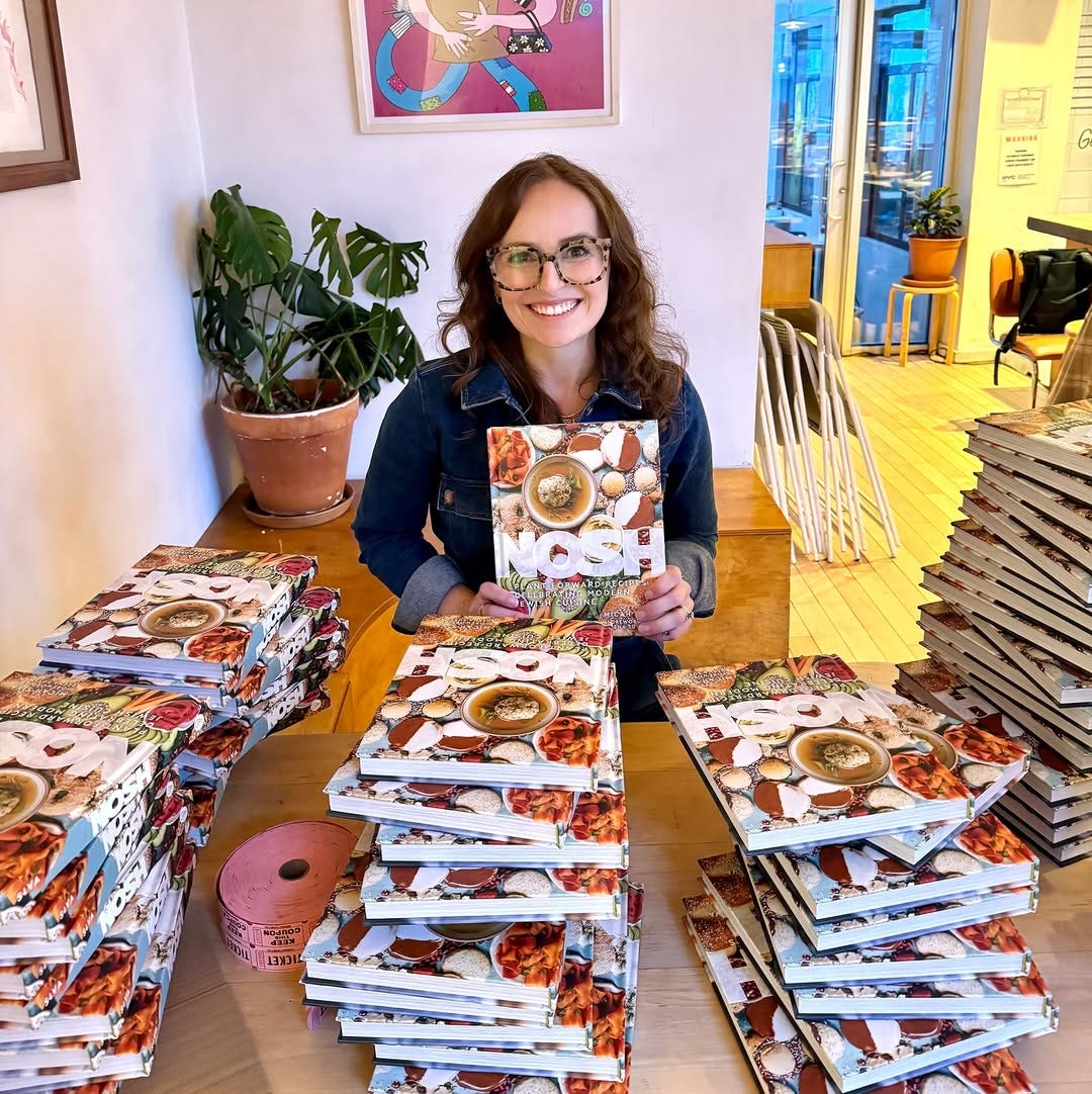 Micah Siva sitting with piles of her cookbook "Nosh" surrounding her Micah Siva sitting with piles of her cookbook "Nosh" surrounding her