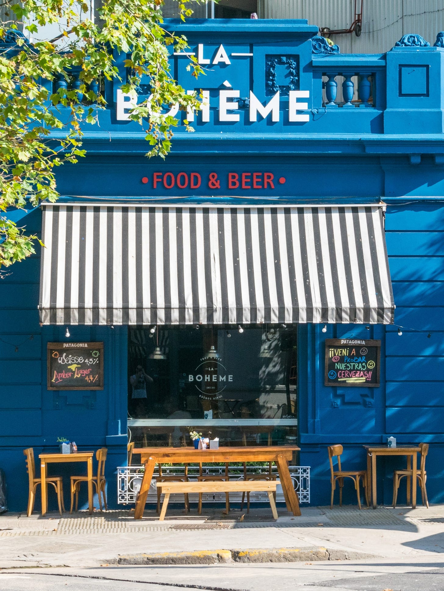 Three wooden tables, benches, and chairs sit outside in the sun next to the blue wall of a restaurant. The restaurant has a white-and-grey striped awning hanging over the seating area to provide shade. Three wooden tables, benches, and chairs sit outside in the sun next to the blue wall of a restaurant. The restaurant has a white-and-grey striped awning hanging over the seating area to provide shade.