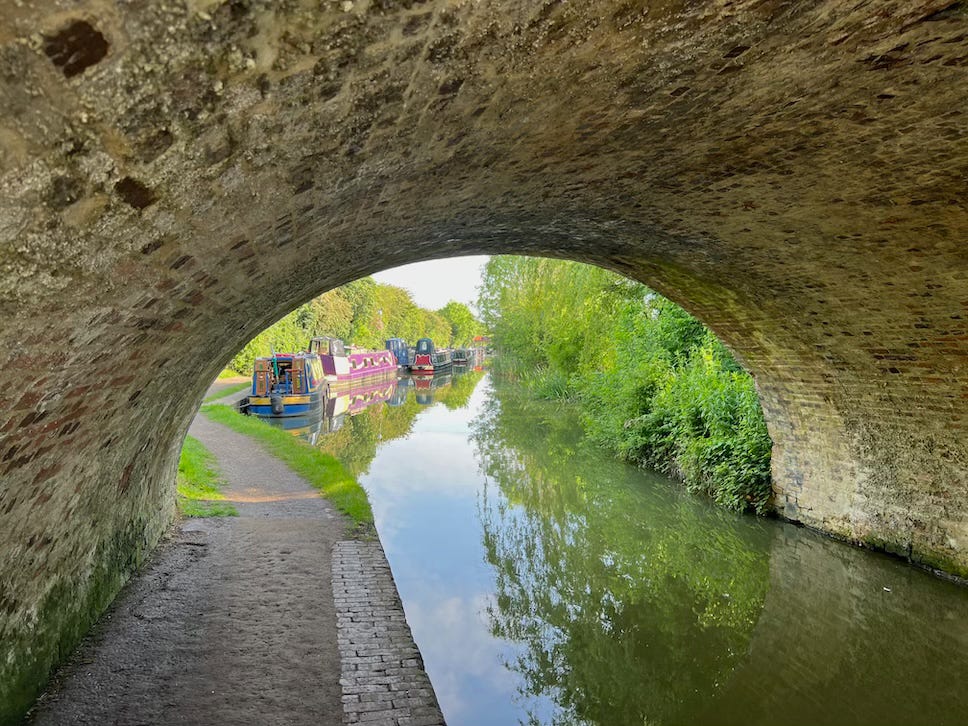 View from beneath a brick canal bridge looking out onto a peaceful canal scene. Several colourful narrowboats are moored along the left bank, with reflections visible in the calm water. A narrow towpath runs alongside the canal, curving slightly out of sight. Lush green vegetation lines both sides of the waterway, and the scene is bathed in soft, natural daylight.