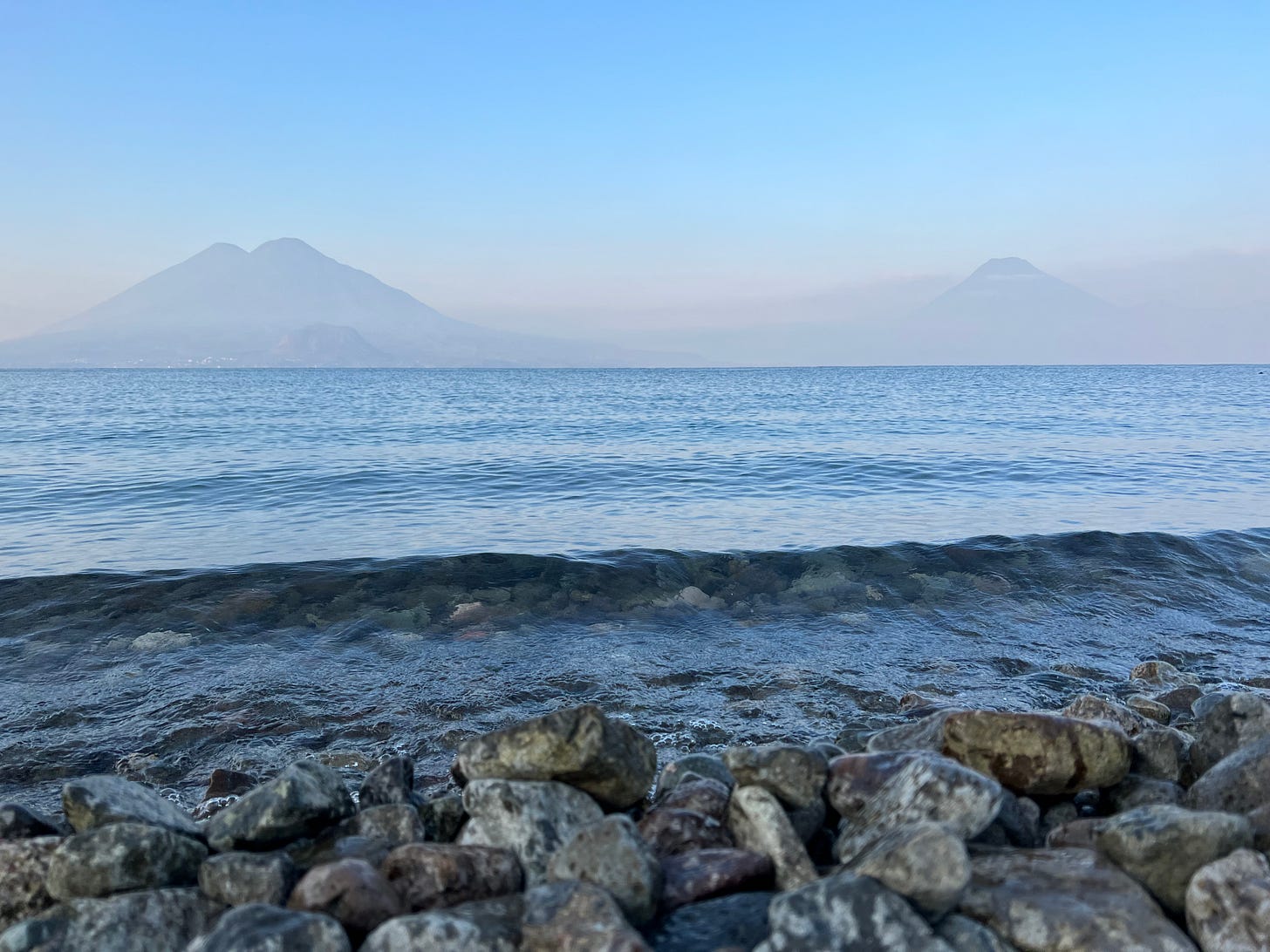a lake in the morning with two volcanos shrouded by haze in the background set against a blue sky