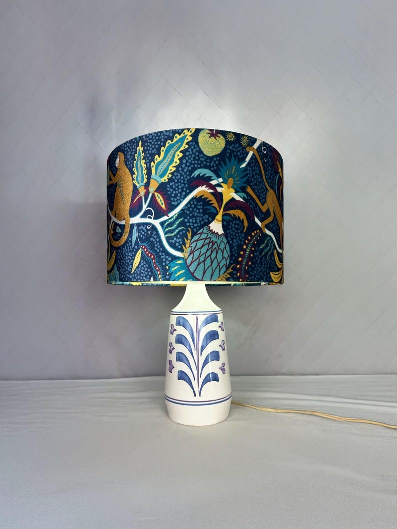 May include: A table lamp with a patterned lampshade and a white ceramic base. The lampshade features a dark blue background with a jungle-themed design, including monkeys and tropical plants. The base has a blue and purple floral pattern.