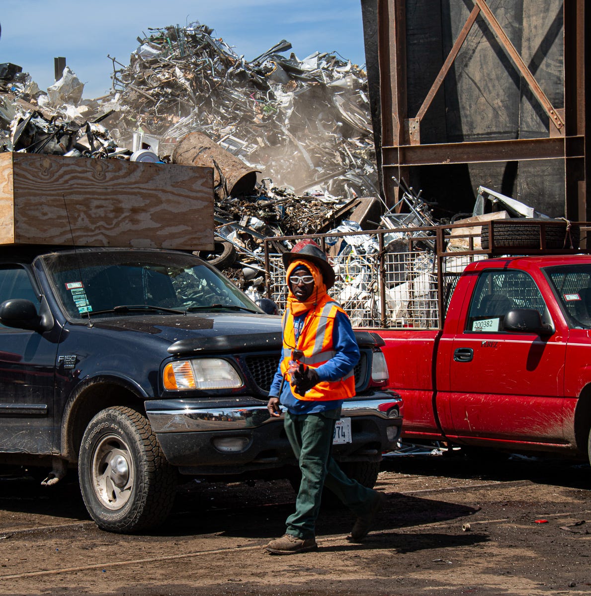 Man in orange safety vest and helmet walking past black and red pickup trucks in a scrapyard, with metal pile in background.