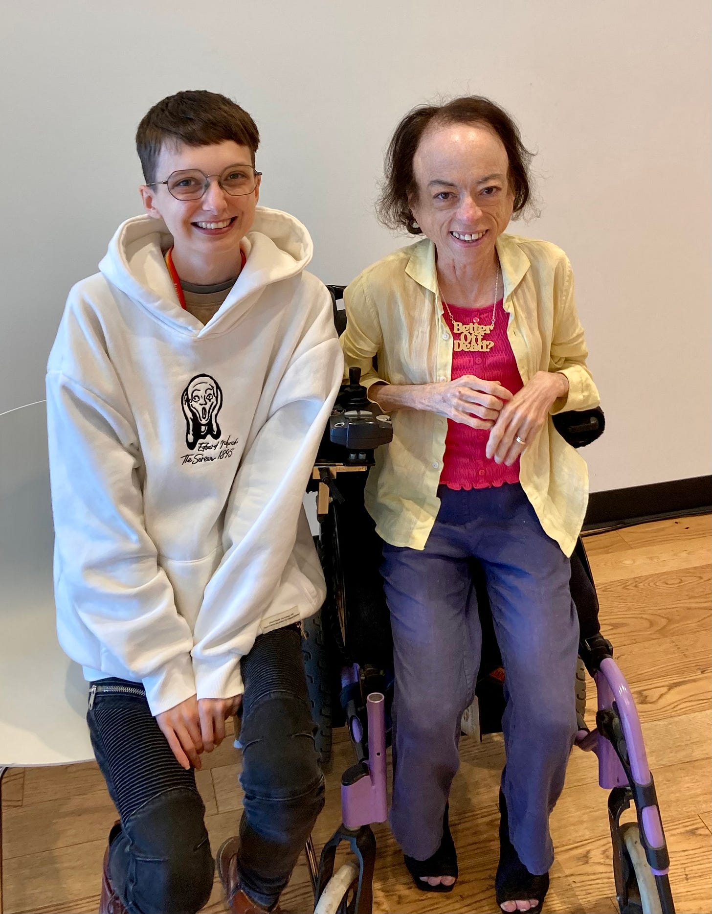 Beck in a hoodie smiling widely next to Liz Carr, a white woman in a powered wheelchair, also smiling