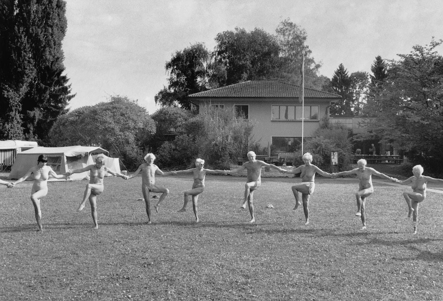 Black and white photograph of eight nude and partially nude older adults standing in a line on a grass lawn, holding hands and lifting one leg in unison. A two-story building and trees are visible in the background, along with a tent and caravan to the left.