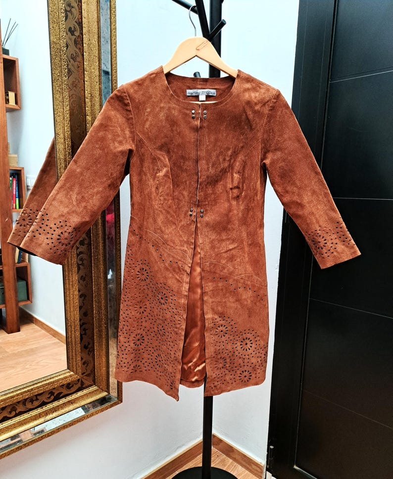 May include: A brown suede jacket with cutouts. The jacket has a round neckline and long sleeves. The jacket is on a hanger and has a zipper closure.