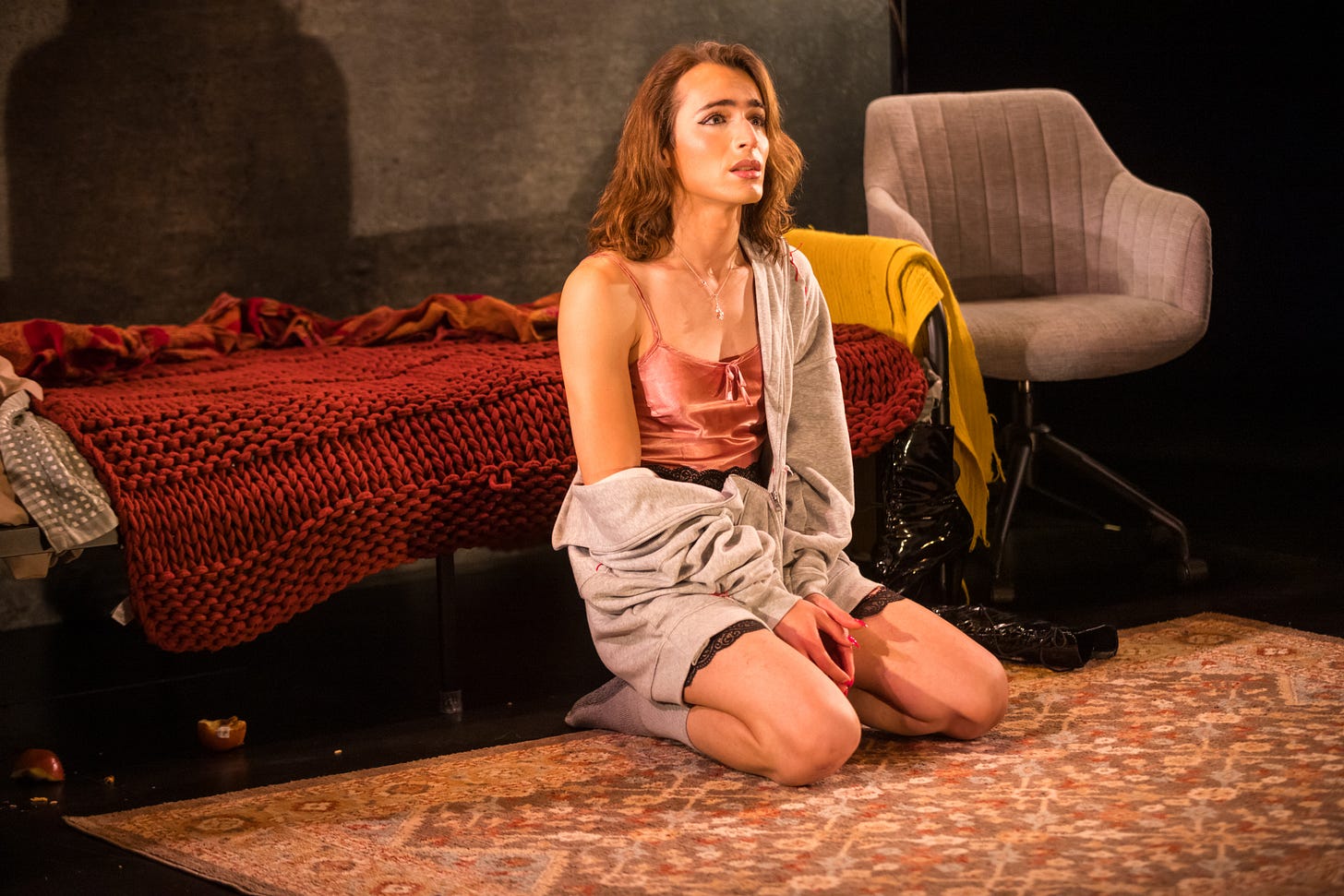 A brunette woman dressed in lingerie and baggy clothes kneels on the carpeted floor of a bedroom