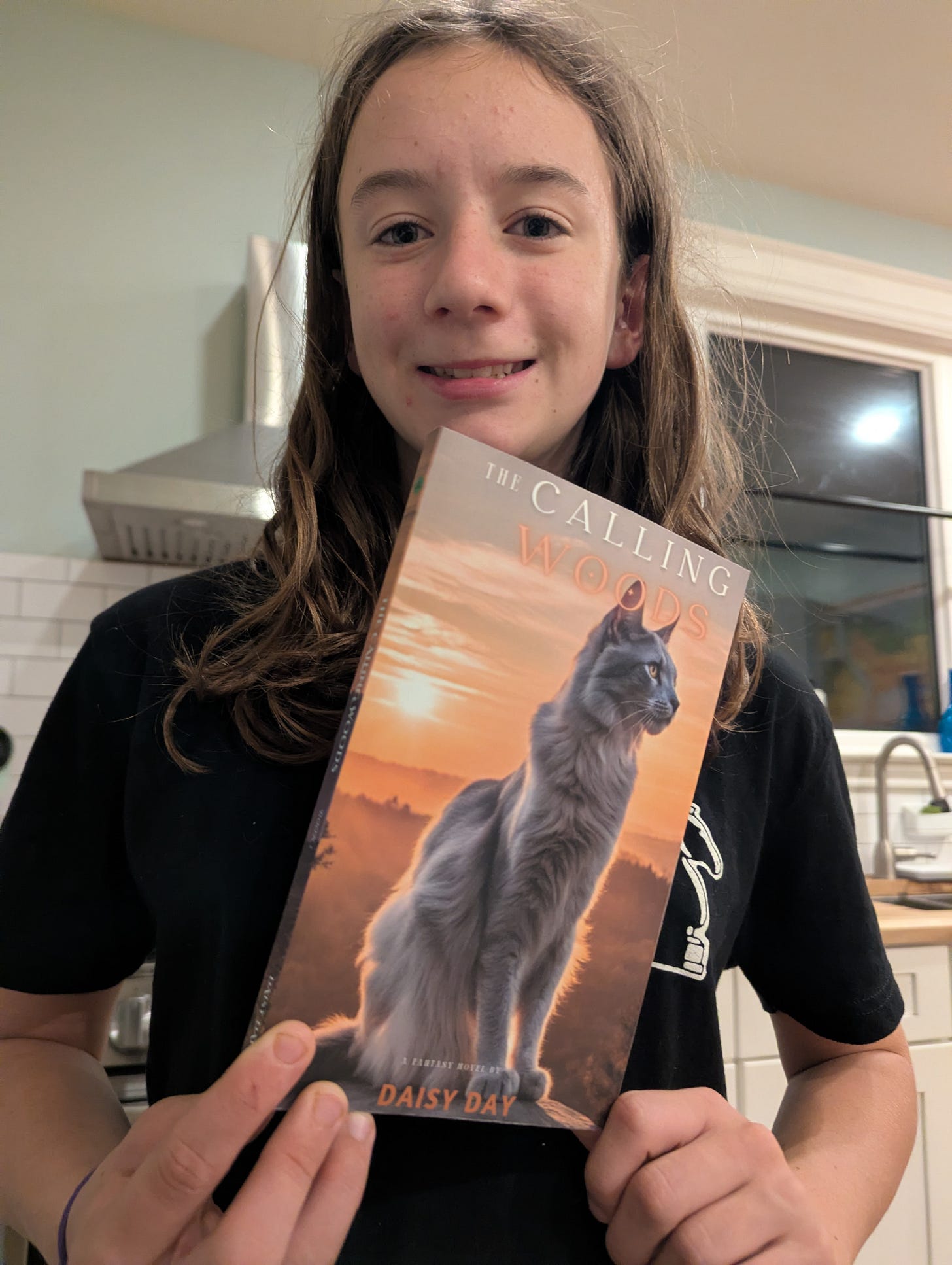 Daisy proudly holds her book. The book has a cat on the cover and is called "The Calling Woods."