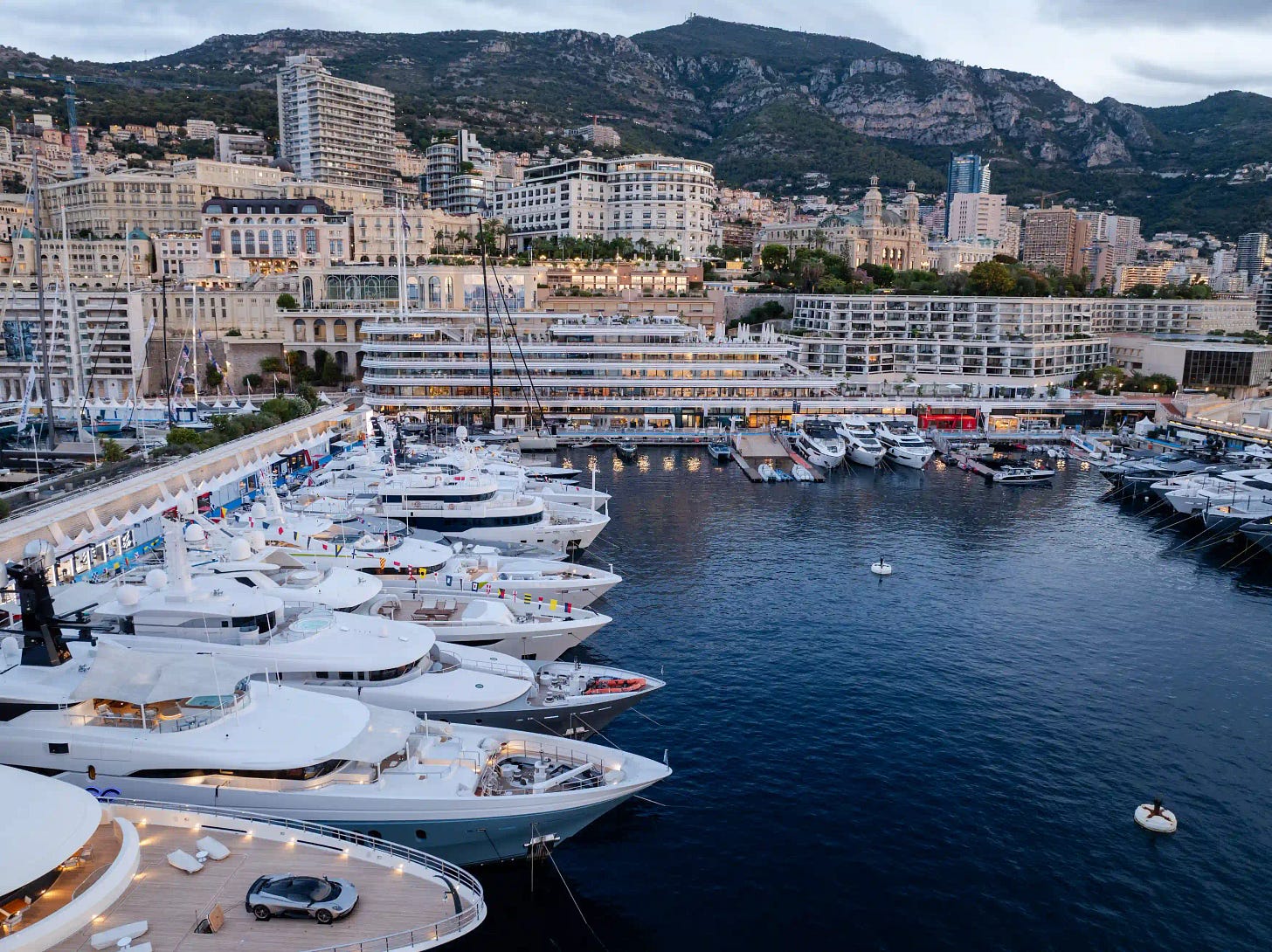 Aerial view of gleaming superyachts moored in Monaco’s Port Hercule, framed by the city’s elegant skyline and mountains.