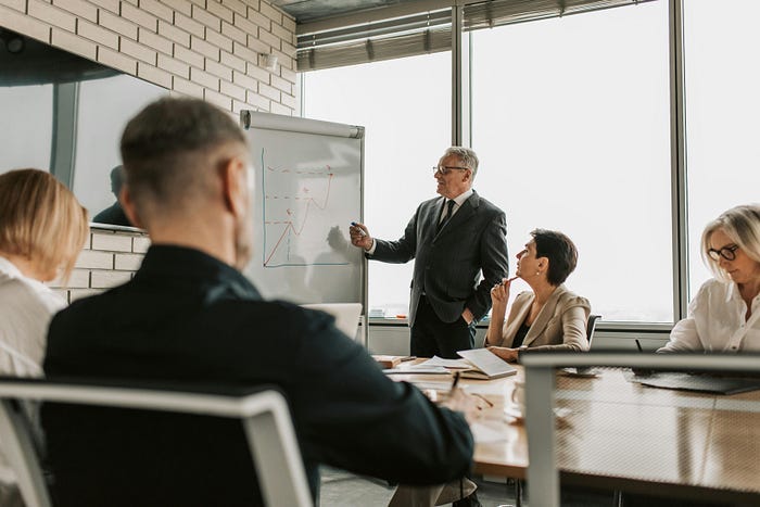A man in a business suit at a whiteboard presenting something, while the rest of the team in business formal attire watches. A man in a business suit at a whiteboard presenting something, while the rest of the team in business formal attire watches.