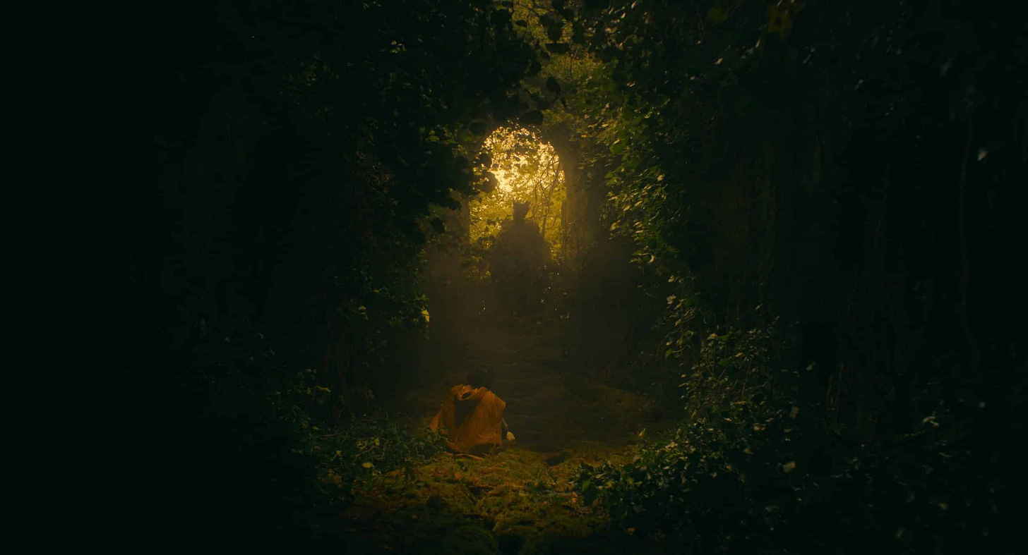 Sir Gawain (Dev Patel) bows before the titular knight (Ralph Ineson) in the overgrowth of a small chapel, as golden light spills in.