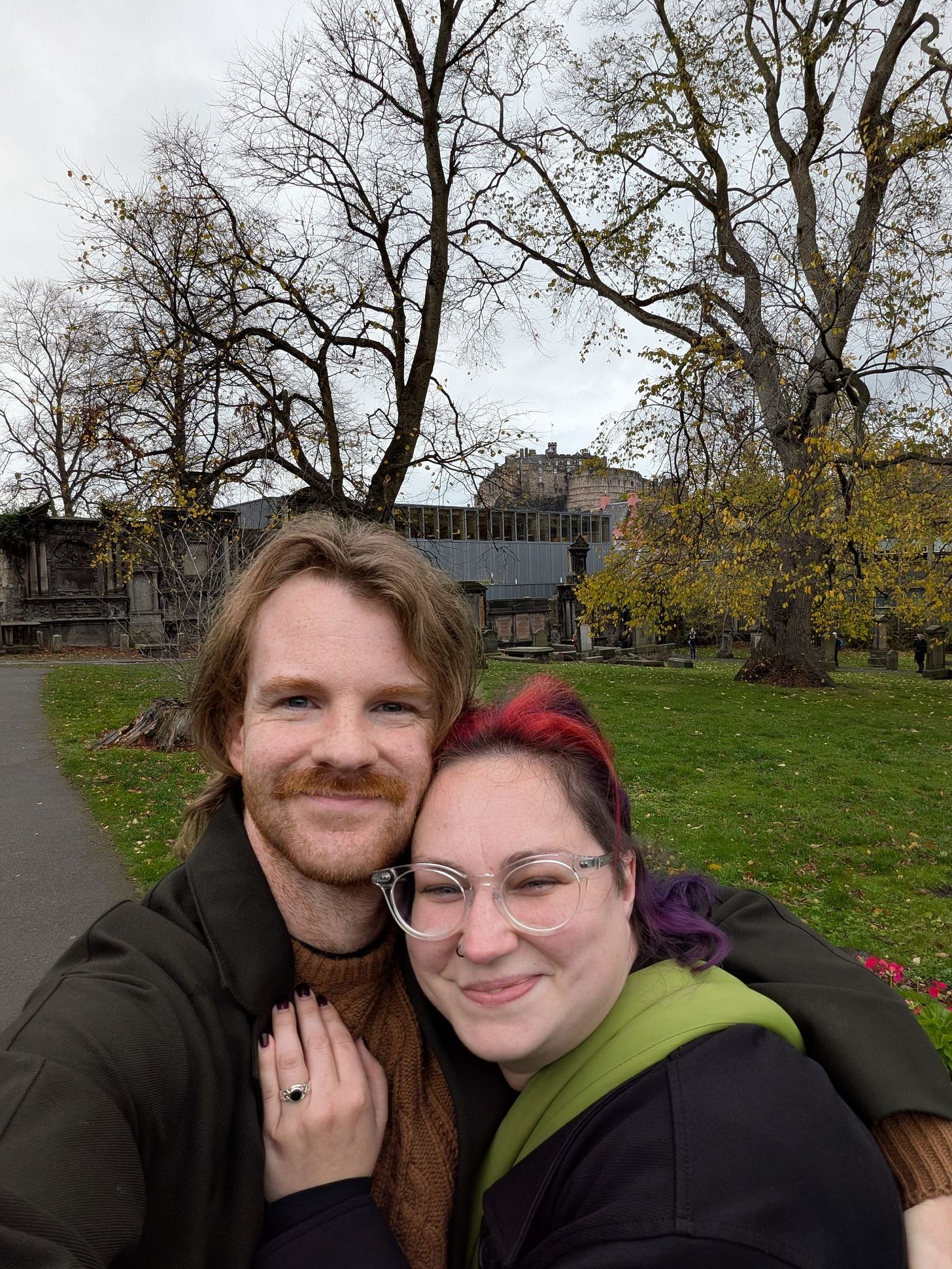 Daire, a white man with ginger hair and moustache stands next to Michelle, a white woman with red and purple hair. Her hand is on his chest, with a ring visible. Behind them is Edinburgh Castle.