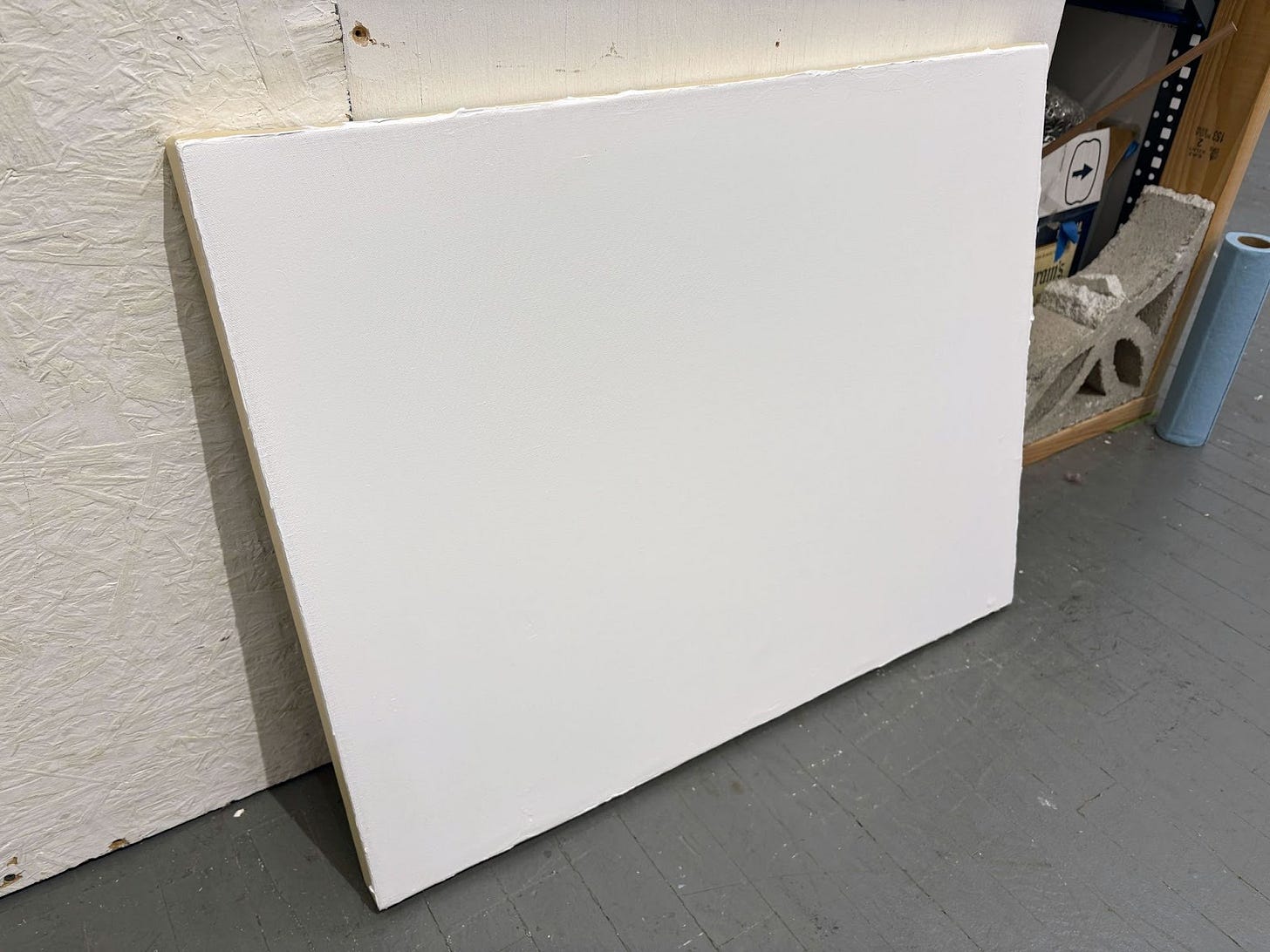 A large white canvas with ground leans against painted wood walls of an art studio.