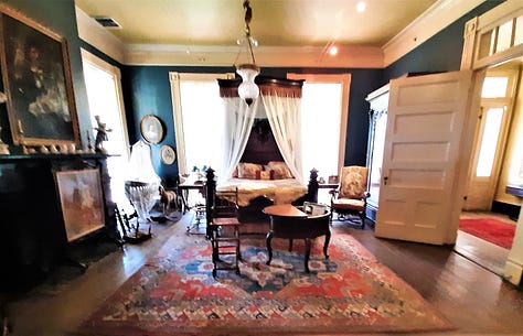 The interior of Houmas House is stately and beautiful with a wide center hall with large portraits on the wall. The second photo shows a fireplace and artwork; the third photo shows a desk in a stately room with a portrait in an ornate frame. The last photos are of bedrooms and a curving staircase.